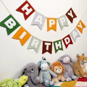 Può includere: Una ghirlanda in tessuto con bandierine a coda di rondine in rosso, arancione, verde e motivi a righe, che compongono la scritta HAPPY BIRTHDAY in lettere bianche. La ghirlanda è appesa sopra una collezione di peluche, tra cui una tartaruga.