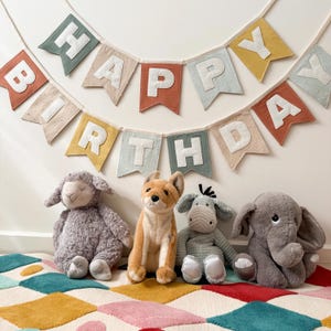 Può includere: Uno striscione colorato di "HAPPY BIRTHDAY" è appeso sopra quattro peluche: una pecora grigia, una volpe marrone, un asino grigio e un elefante grigio. I giocattoli sono su un tappeto fantasia con vari colori.