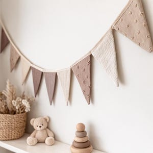 Puede incluir: Escena de decoración de guardería en tonos neutros. Guirnalda de banderas triangulares en tonos beige y marrón. Un osito de peluche y un juguete apilable de madera sobre una estantería blanca. Cesta de mimbre con flores secas.