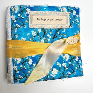 May include: A square fabric-covered journal featuring a bright blue background with white floral branches. A rectangular beige fabric label with the text "be happy and create" is stitched on the front. A yellow ribbon is tied around the center.