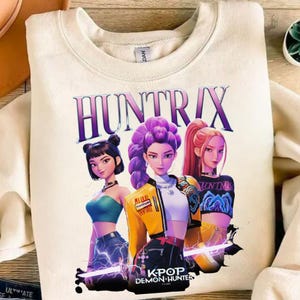 May include: A cream-coloured sweatshirt with a graphic of three anime-style women with colourful hair. The text "HUNTRIX" is above the figures, with "K-POP DEMON HUNTER" below. The women are in stylish outfits with lightning effects.