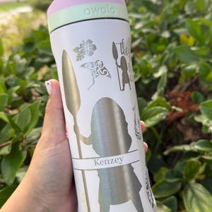 May include: White Owala water bottle with a pink and green lid. The bottle features silver graphics of a woman with a spear, floral designs, and the name "Kenzey". The text "the...is calling" is also visible. The bottle is held in a hand.