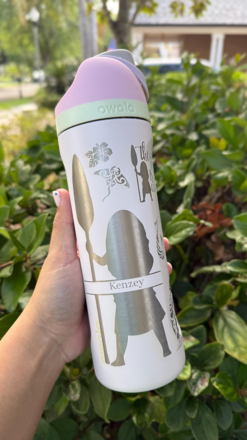 May include: White Owala water bottle with a pink and green lid. The bottle features silver graphics of a woman with a spear, floral designs, and the name "Kenzey". The text "the...is calling" is also visible. The bottle is held in a hand.