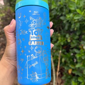 May include: Blue water bottle featuring Toy Story characters and text. The bottle has a green lid and a silver design with the words "Buzz Lightyear", "Toy Story", "Carter", and "Sunnyside".