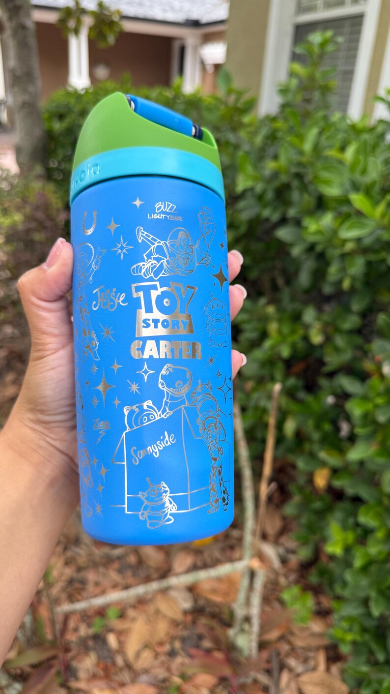 May include: Blue water bottle featuring Toy Story characters and text. The bottle has a green lid and a silver design with the words "Buzz Lightyear", "Toy Story", "Carter", and "Sunnyside".