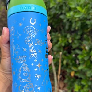 May include: A blue Owala water bottle featuring gold illustrations of characters from the Toy Story franchise, including Jessie, Bullseye, Mr. Potato Head, and Zurg. The bottle has a green and blue lid and a black handle.