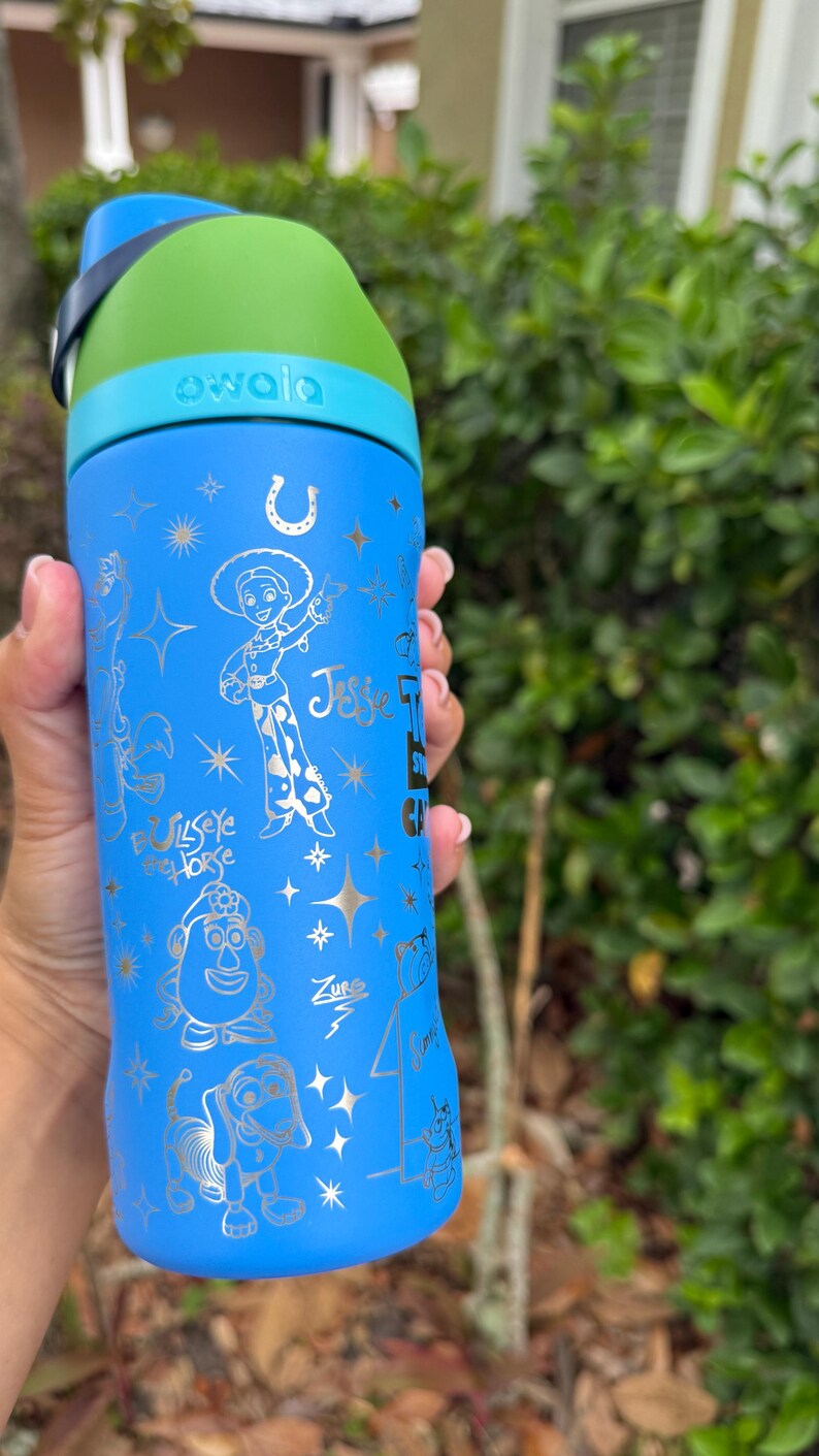 May include: A blue Owala water bottle featuring gold illustrations of characters from the Toy Story franchise, including Jessie, Bullseye, Mr. Potato Head, and Zurg. The bottle has a green and blue lid and a black handle.