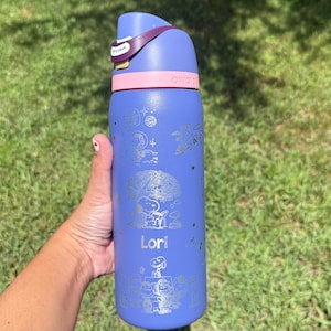 May include: A blue water bottle with a pink band and a purple strap. The bottle is decorated with silver Snoopy and Woodstock designs and the name "Lori". The bottle is held against a background of green grass.