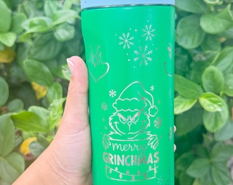 Puede incluir: Una botella de agua de color verde brillante con un gráfico plateado de un personaje con gorro de Papá Noel, copos de nieve y el texto "merry GRINCHMAS". La botella tiene un cuello azul claro, una tapa blanca con "Lift to Unlock" y un tapón verde lima.