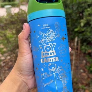 May include: Blue water bottle featuring Buzz Lightyear and other Toy Story characters. The bottle has a green and blue lid and the text "Toy Story" and "Sunnyside" in gold lettering. The bottle is approximately 8 inches tall.