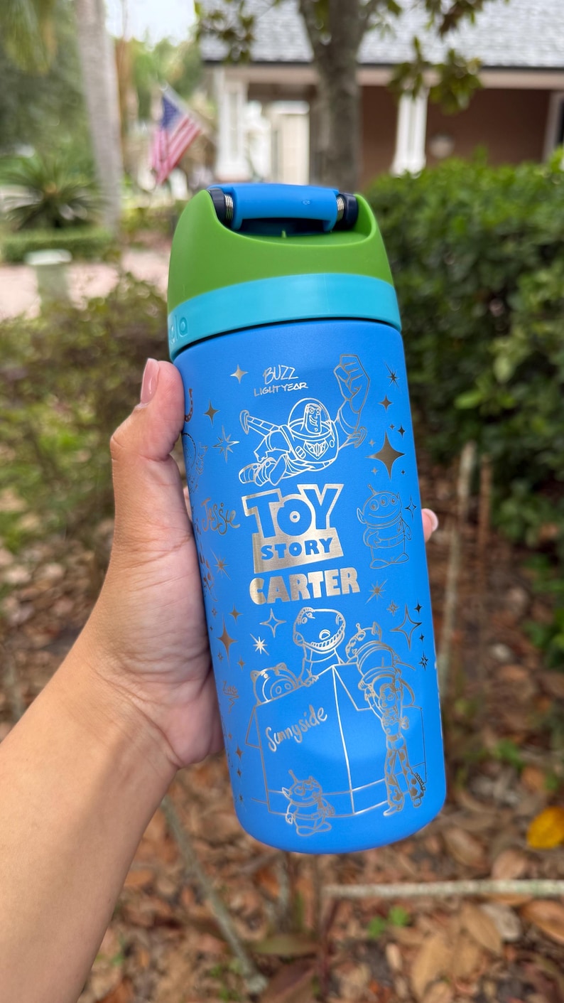 May include: Blue water bottle featuring Buzz Lightyear and other Toy Story characters. The bottle has a green and blue lid and the text "Toy Story" and "Sunnyside" in gold lettering. The bottle is approximately 8 inches tall.