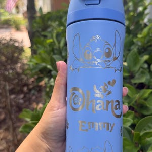 Könnte beinhalten: Eine hellblaue Wasserflasche mit einem goldfarbenen Cartoon-Charakter-Design und dem Wort "Ohana" mit dem Namen "Emmy". Die Flasche hat einen blauen Deckel mit dem Text "Lift to Unlock".