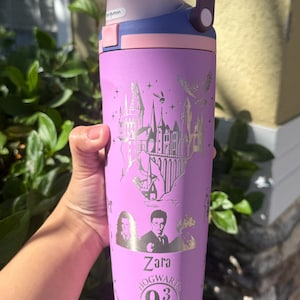 May include: A purple water bottle with a handle and a flip-top lid. The bottle features a silver design of a castle, characters, and the text "Zara Hogwarts 9 3/4 Express". The bottle is held against a background of green foliage.