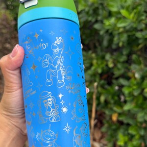 May include: Blue water bottle with a green and blue lid. The bottle features gold illustrations of characters from a popular animated movie. The text "Lift to Unlock" is visible on the lid.
