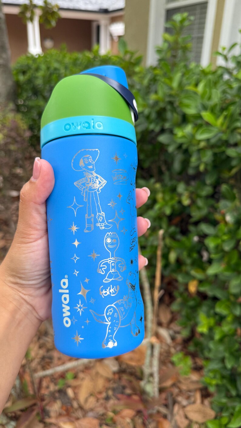 May include: Blue Owala water bottle with a green lid and a blue handle. The bottle features silver illustrations of characters from a popular animated film. The word "owala" is printed on the bottle.