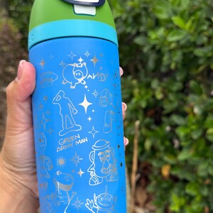 May include: Blue water bottle with a green and blue lid. The bottle features gold illustrations of characters and phrases from the movie Toy Story. The lid has the text "Lift to Unlock".
