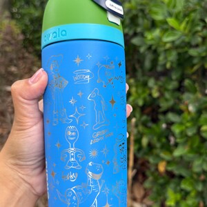 May include: A blue Owala water bottle with a green top and blue handle. The bottle features gold illustrations of characters from the movie Toy Story, including Woody, Buzz Lightyear, and Rex. The bottle has the word "owala" printed on it.