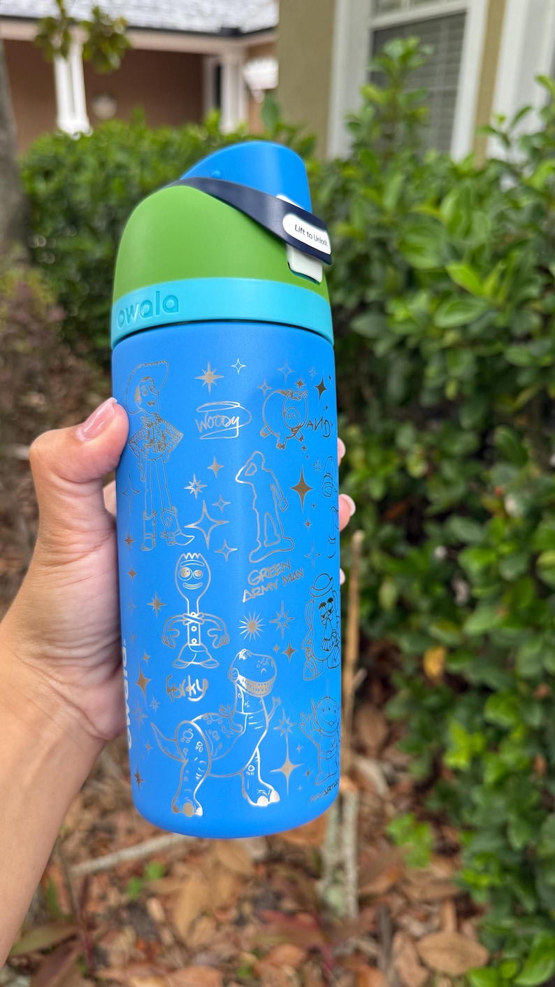 May include: A blue Owala water bottle with a green top and blue handle. The bottle features gold illustrations of characters from the movie Toy Story, including Woody, Buzz Lightyear, and Rex. The bottle has the word "owala" printed on it.