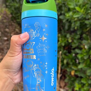 May include: A blue Owala water bottle featuring Toy Story characters. The bottle has a blue, green, and teal lid and handle. The bottle has the words "Toy Story" and "Carter" printed on it.