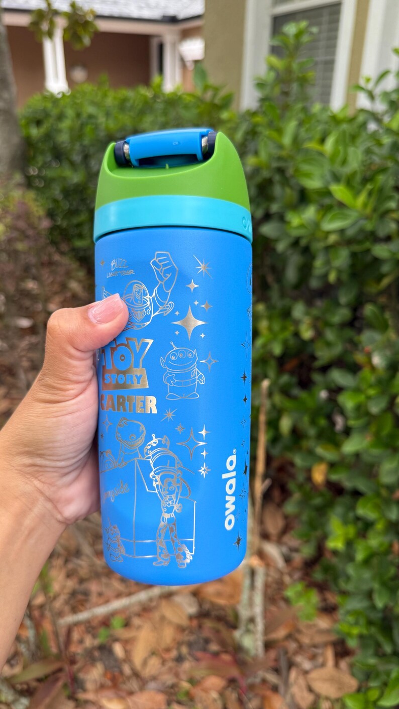 May include: A blue Owala water bottle featuring Toy Story characters. The bottle has a blue, green, and teal lid and handle. The bottle has the words "Toy Story" and "Carter" printed on it.