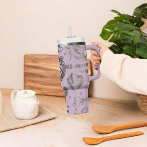 May include: A lavender insulated tumbler with a handle, a white lid, and a silver straw. The tumbler is decorated with silver illustrations and the text "Name xog". The tumbler is designed to keep beverages cold or hot.
