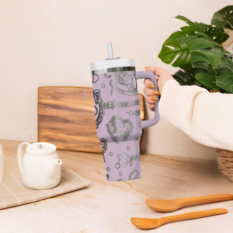 May include: A lavender insulated tumbler with a handle, a white lid, and a silver straw. The tumbler is decorated with silver illustrations and the text "Name xog". The tumbler is designed to keep beverages cold or hot.