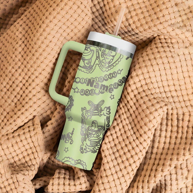May include: A light green tumbler with a white lid and handle, featuring a decorative design and the word "Name". The tumbler includes a straw and is set against a textured, tan fabric.