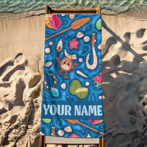Pode incluir: Uma toalha de praia azul com um design tropical com personagens de desenhos animados, conchas e um anzol. A toalha tem o texto "YOUR NAME" em branco na parte inferior. A toalha est&aacute; em uma cadeira de praia de madeira na areia.