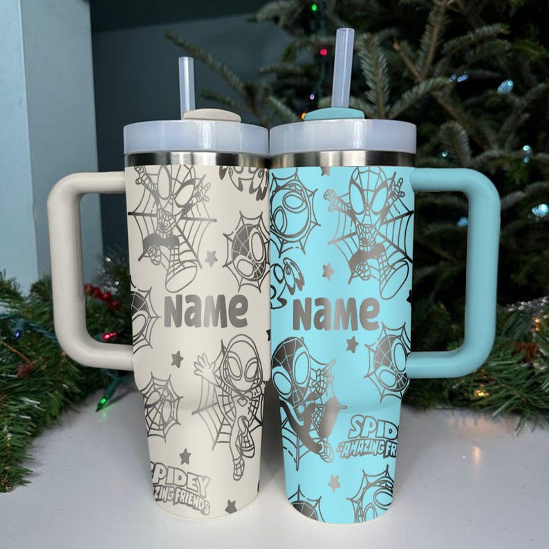 Puede incluir: Dos vasos aislados con asas, uno blanquecino y otro azul claro. Cada uno presenta un dise&ntilde;o de Spider-Man con una telara&ntilde;a, estrellas y la palabra "Name". El vaso azul claro tambi&eacute;n tiene el texto "Spidey and Amazing Friends".