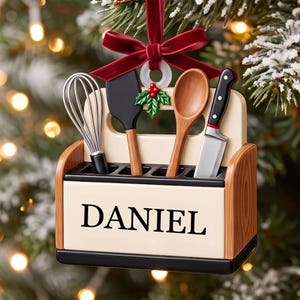 May include: A decorative Christmas ornament shaped like a kitchen utensil caddy with the name DANIEL printed in black. It holds a miniature whisk, spatula, wooden spoon, and knife. The caddy is topped with a red velvet bow and holly leaves.