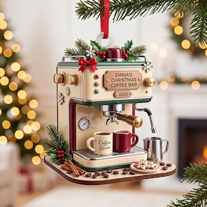 May include: A cream and dark red espresso machine Christmas ornament featuring a sign that reads EMMA'S CHRISTMAS COFFEE BAR 2026. It includes two small mugs, one labeled Coffee Lover, a metal pitcher, cinnamon sticks, and cookies on a base.
