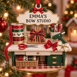 May include: A decorative Christmas ornament shaped like a craft table with a sign reading EMMA'S BOW STUDIO. It features spools of ribbon, a bow maker, red scissors, a green glue gun, two finished bows, and a wicker basket filled with ribbon rolls.