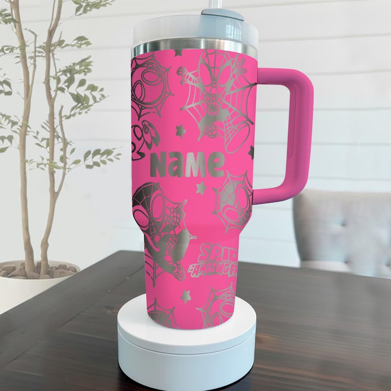 Puede incluir: Un vaso t&eacute;rmico rosa con un dise&ntilde;o plateado de Spider-Man y un asa. El vaso presenta las palabras "NAME" y "Spider-Man" en plata. El vaso est&aacute; sobre un soporte blanco.