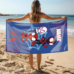 Puede incluir: Una toalla de playa azul con personajes de Spider-Man. La toalla tiene un gran gr&aacute;fico de Spider-Man, Spider-Gwen y Miles Morales. La palabra "NAME" est&aacute; impresa en blanco en el lado derecho de la toalla. La toalla se sostiene en una playa de arena.
