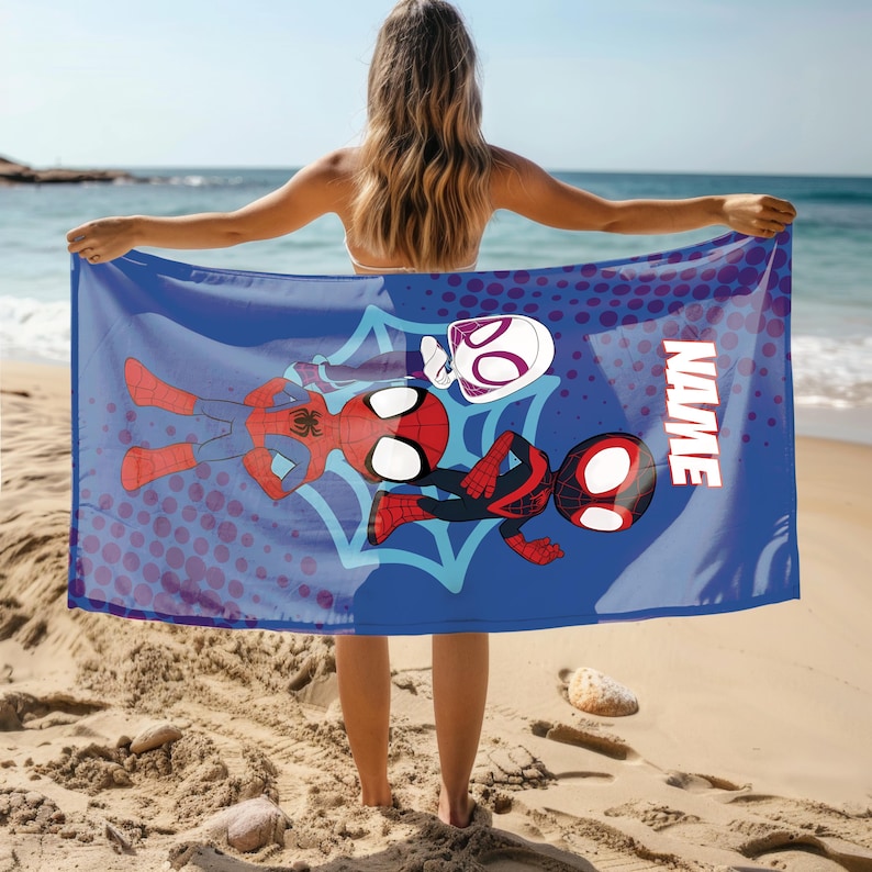Puede incluir: Una toalla de playa azul con personajes de Spider-Man. La toalla tiene un gran gr&aacute;fico de Spider-Man, Spider-Gwen y Miles Morales. La palabra "NAME" est&aacute; impresa en blanco en el lado derecho de la toalla. La toalla se sostiene en una playa de arena.