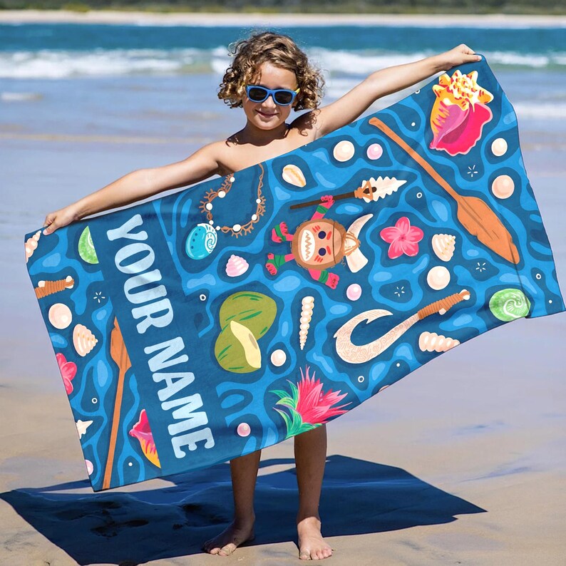 Pode incluir: Uma toalha de praia azul com um design tropical com conchas, cocos e uma personagem de desenho animado. A toalha tem o texto "YOUR NAME" em branco. A toalha &eacute; segurada por uma pessoa numa praia arenosa.