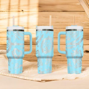 May include: Three light blue insulated tumblers with white lids, handles, and straws. Each tumbler features a silver floral design and the name "Nina" printed on the side. The tumblers are displayed on a white cloth against a wooden background.