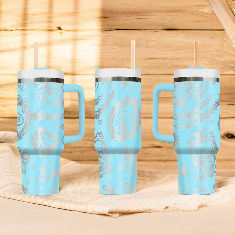 May include: Three light blue insulated tumblers with white lids, handles, and straws. Each tumbler features a silver floral design and the name "Nina" printed on the side. The tumblers are displayed on a white cloth against a wooden background.
