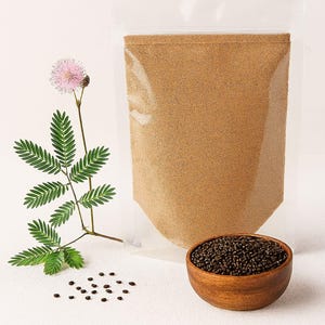 Può includere: Un sacchetto di plastica trasparente pieno di polvere marrone, una piccola ciotola di legno piena di semi neri e un rametto di foglie verdi e un fiore rosa. Gli oggetti sono disposti su una superficie bianca.