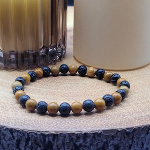 May include: A bracelet made of black lava rock and light brown wooden beads. The bracelet is displayed on a wooden surface, with a candle and a decorative glass jar in the background. The bracelet is a neutral accessory, suitable for any outfit.