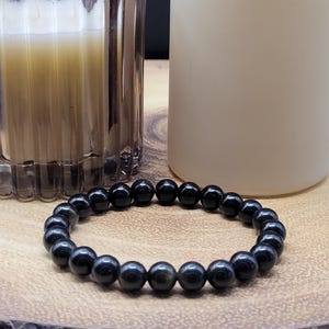 May include: A circular stretch bracelet composed of smooth, polished, spherical black beads. The beads exhibit a subtle, reflective sheen across their dark surfaces, arranged in a continuous loop to form a simple, uniform band of dark stone.