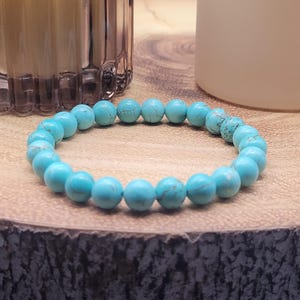 May include: A turquoise beaded bracelet. The bracelet features round, light blue beads with natural colour variations. The beads are strung together, forming a circular shape. The bracelet is displayed on a wooden surface.