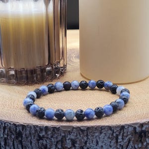 May include: A beaded bracelet featuring an alternating pattern of smooth, mottled blue and white stone beads and porous, textured black lava beads. The circular jewellery piece is strung on an elastic cord, showcasing a contrast between the polished blue spheres and the rough, dark volcanic rock beads.