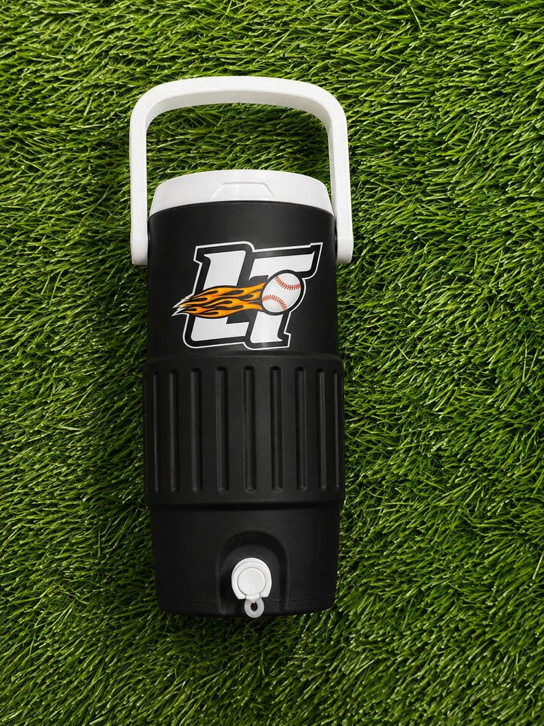 May include: Black and white insulated water cooler with a white handle. The cooler features a baseball logo with a baseball and flames. The cooler has a white spigot and is on a green surface.
