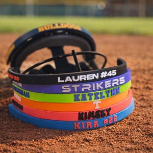 Könnte beinhalten: Ein Baseballhelm mit einem Stapel bunter Stirnbänder. Die Stirnbänder sind schwarz, lila, grün, orange und blau, jedes mit einem Namen und einer Nummer in einer Kontrastfarbe. Der Helm ist marineblau und gelb.