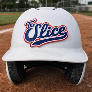 May include: A white baseball helmet with the text "The Slice" in red and blue lettering. The helmet is sitting on a brown surface, likely a baseball field. The helmet has black padding around the ears.
