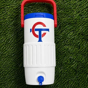 May include: White water cooler with a red handle and a blue and red logo. The cooler has a blue lid and a blue spigot. The cooler is sitting on a green surface.