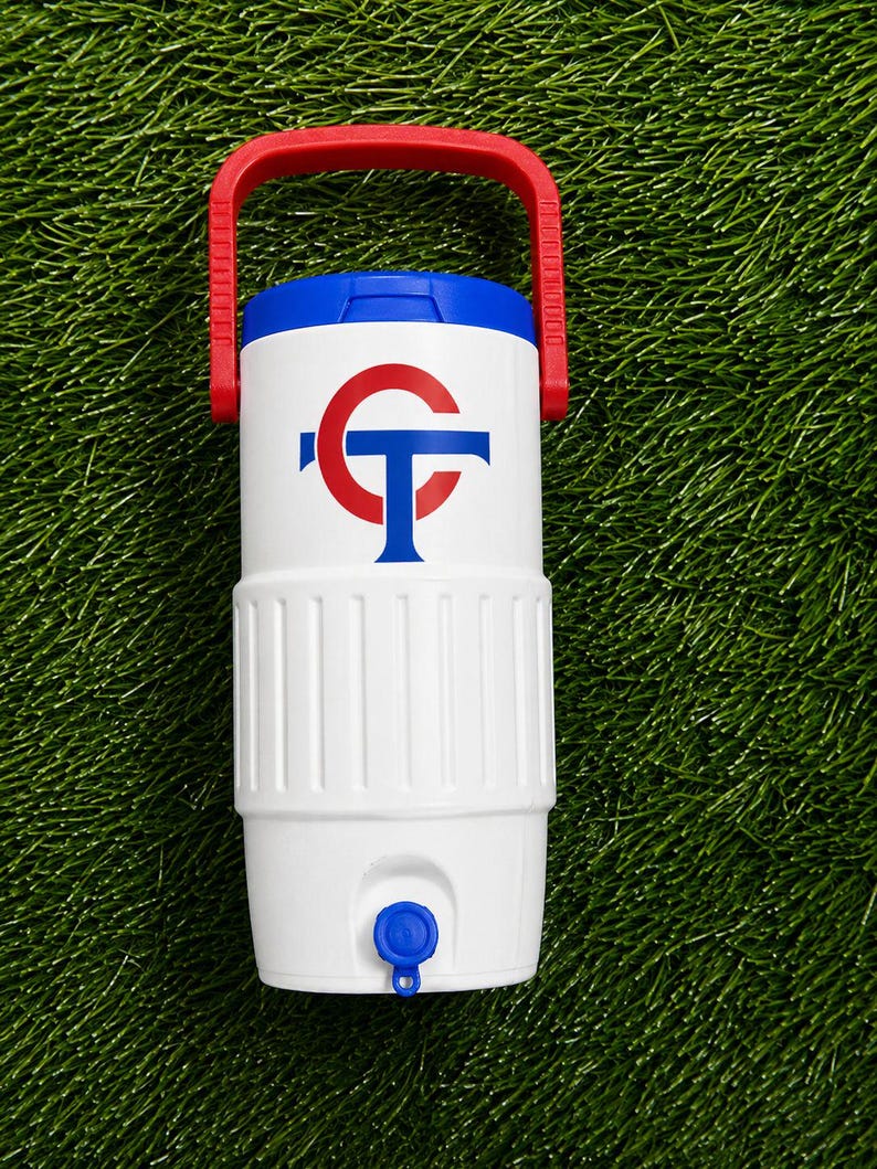 May include: White water cooler with a red handle and a blue and red logo. The cooler has a blue lid and a blue spigot. The cooler is sitting on a green surface.