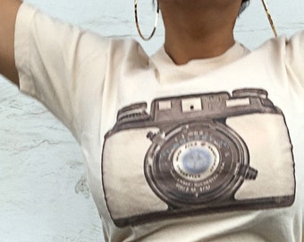 May include: A cream-colored t-shirt featuring a vintage camera graphic. The camera design is in shades of brown and gray. The person is wearing black jeans. The background is a white wall.