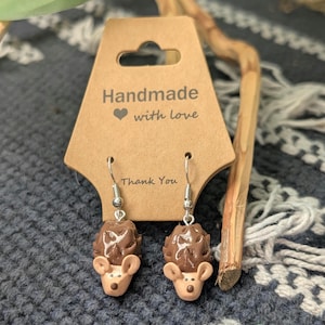 May include: A pair of dangle earrings featuring sculpted brown and tan hedgehog charms with dark brown noses. The earrings are attached to a tan cardboard display card printed with the text Handmade with love and Thank You.
